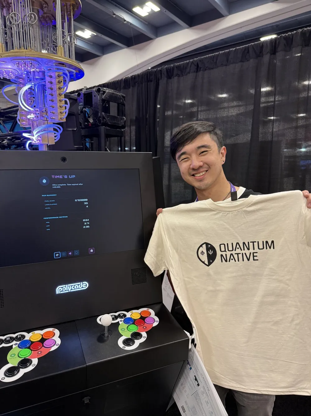 A Quantum Native shirt held beside the Quantum Arcade cabinet, arcade stick and buttons in view, at GDC 2026